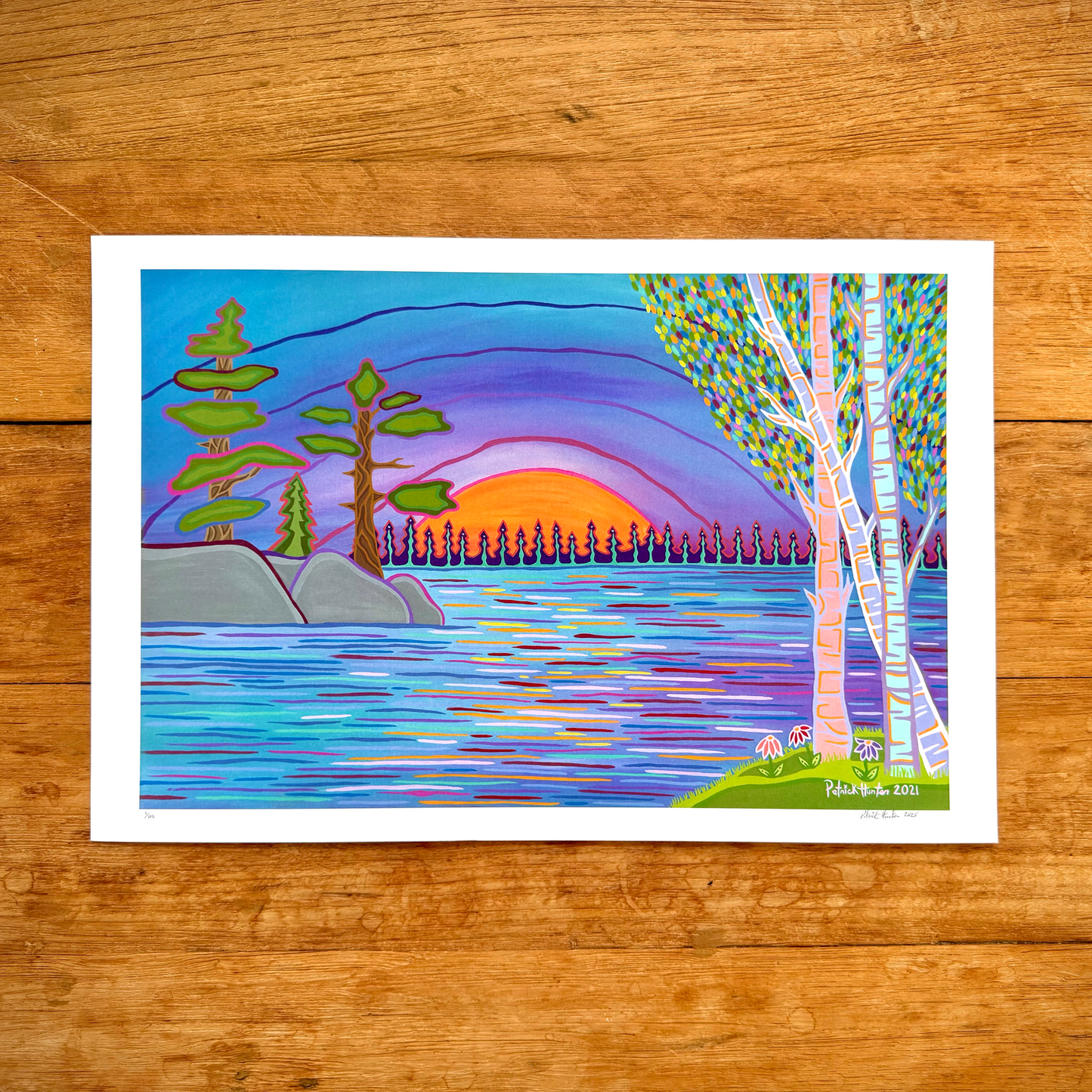 A print of Patrick Hunter's painting, Sunset Bay, a woodland art style landscape of open water with trees and sunset framing. The print rests on a wood grain background.