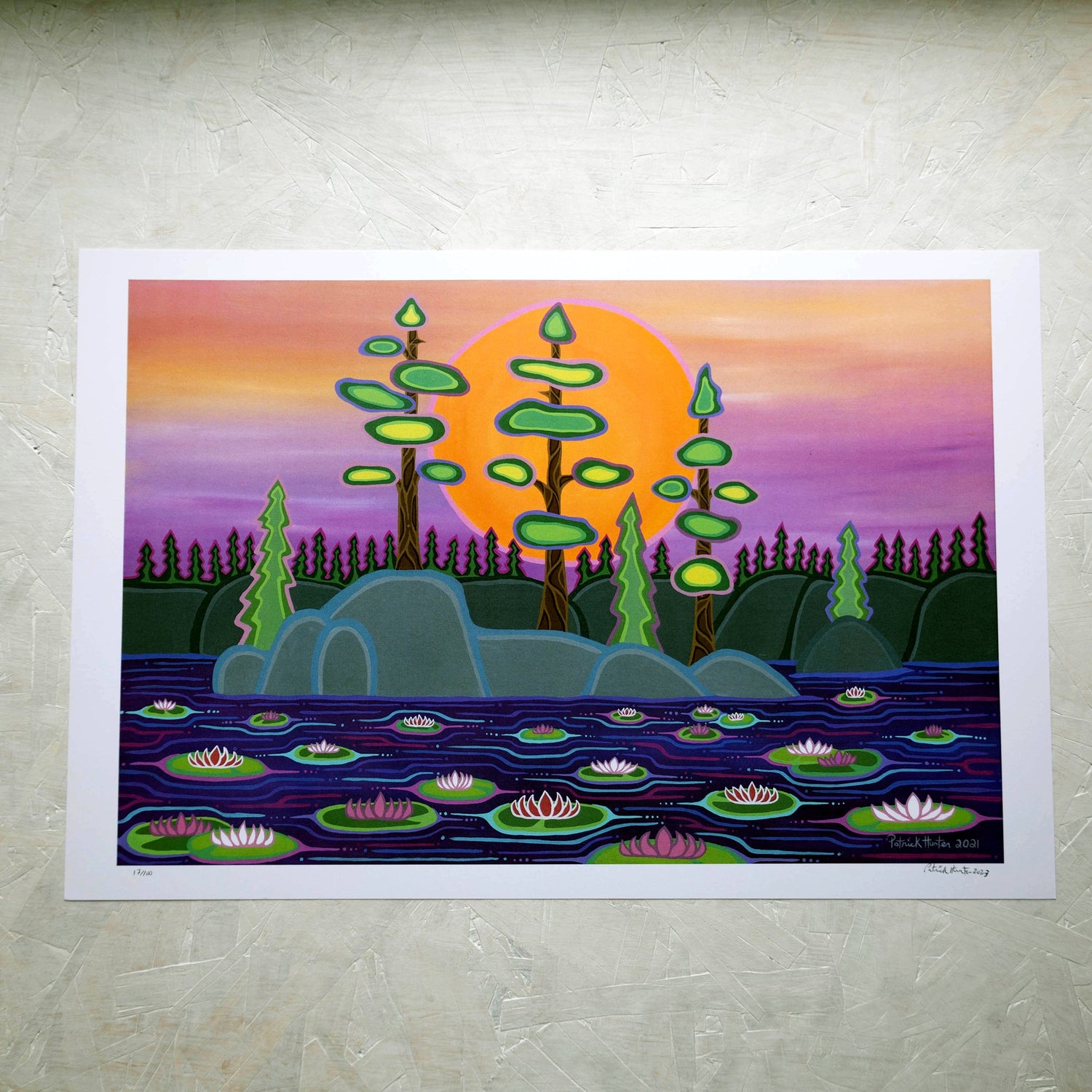 A print of Patrick Hunter's painting of lily pads on water in front of a rock with several trees, against the backdrop of a sun low in the horizon, in woodland art style.