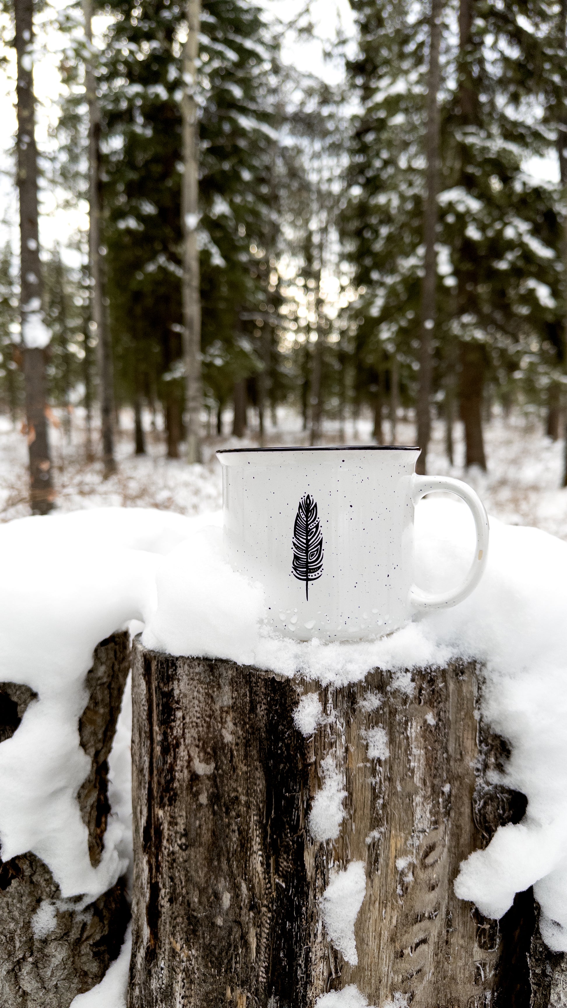 Ceramic mug with speckled finish and black rim, boasting Patrick's eagle feather in woodland art style, rests on a tree stump surrounded by snow. A snowy forest is out of focus in the background.