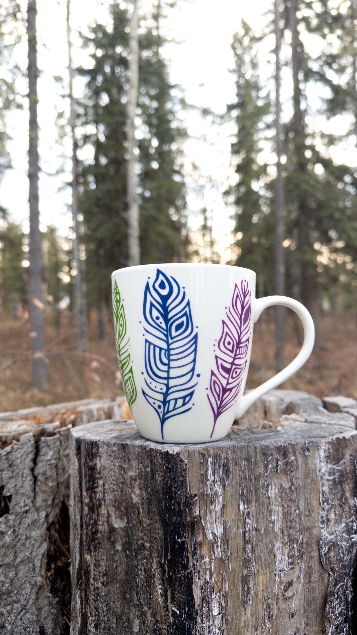 Patrick's Pride Feathers on a tall white mug, resting on a tree stump. The image shows the green, blue, and purple feathers of one side.