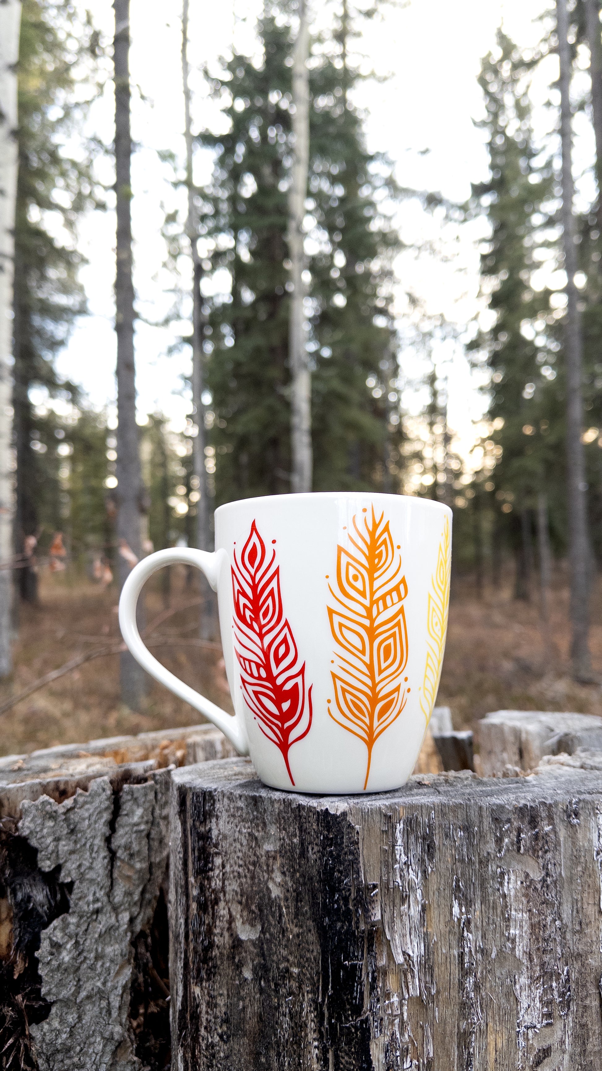 Patrick's Pride Feathers on a tall white mug, resting on a tree stump. The image shows the red, orange, and yellow feathers on one side.