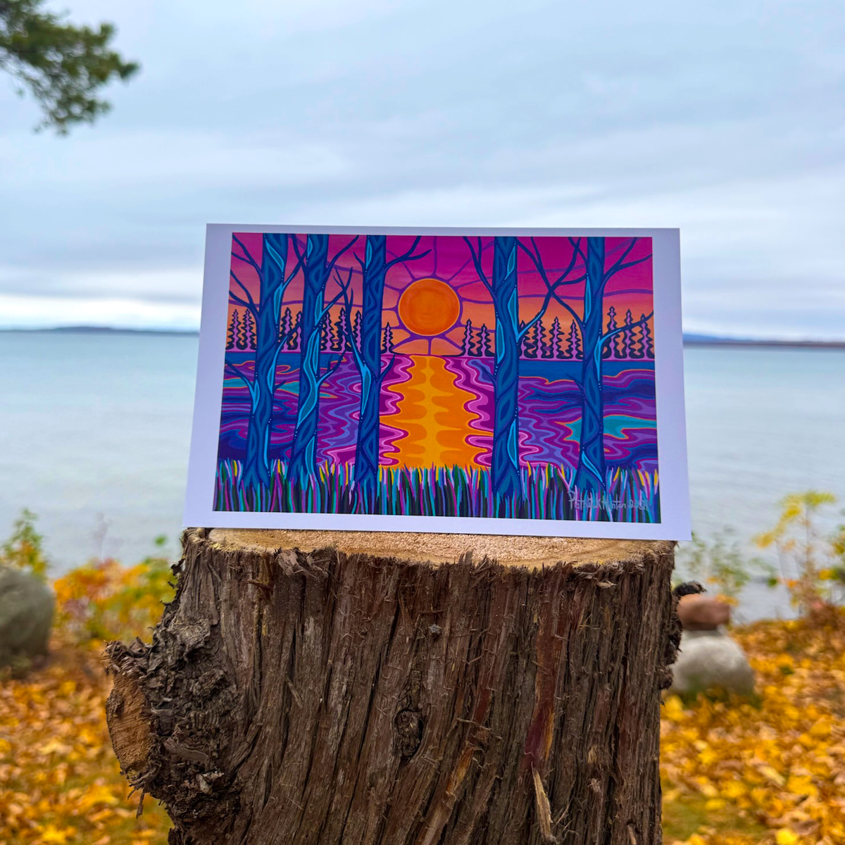 A greeting card with Patrick's woodland art style painting of a sunset through trees, with water and more trees in the background.
