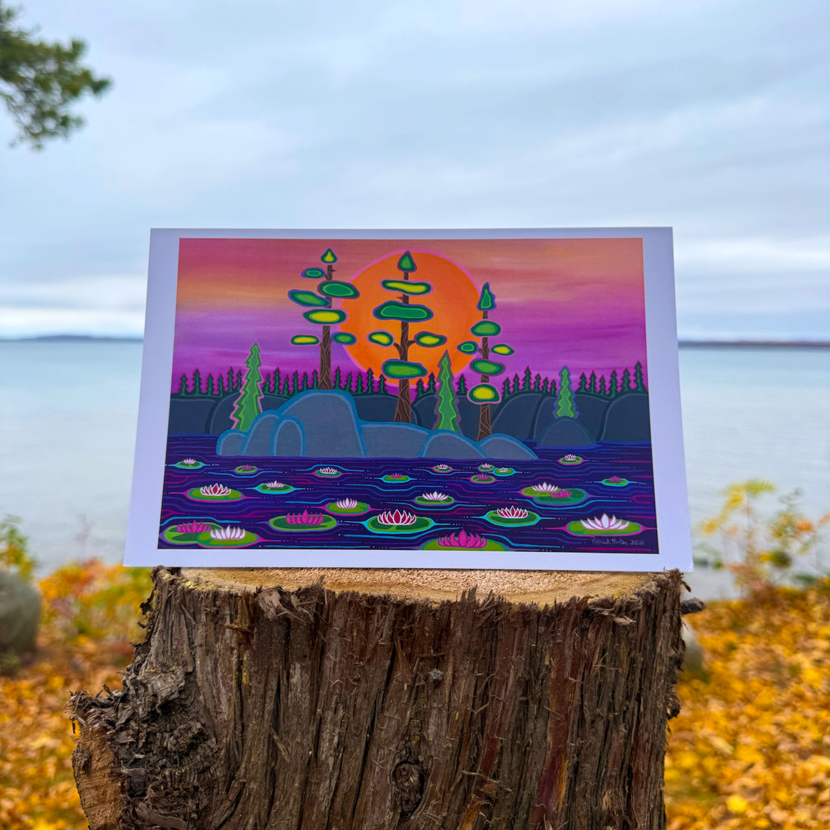 A greeting card with Patrick's woodland art style painting of trees against a sunset, with water lilies in the foreground.