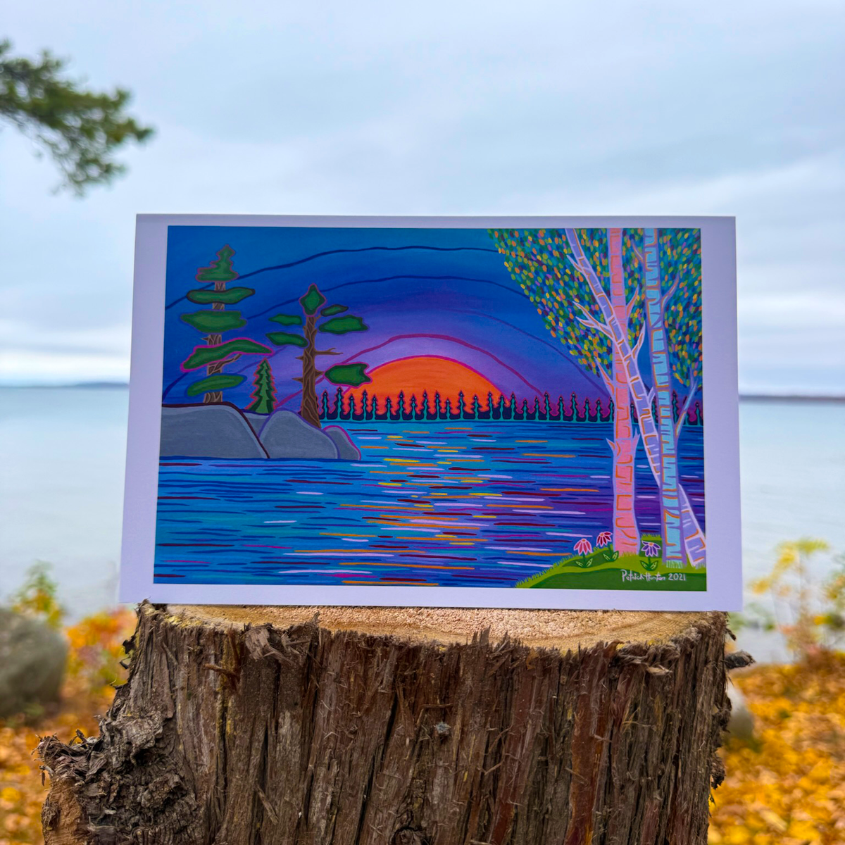 A greeting card of Patrick's woodland art style painting of a sunset over a body of water with trees in the mid and foreground.