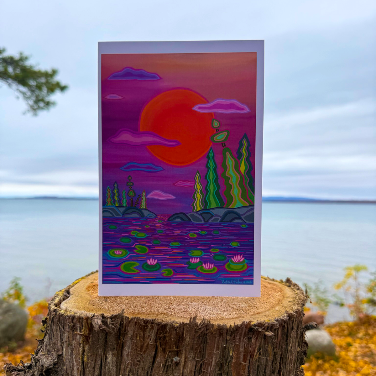 A greeting card of Patrick's woodland art style painting of a sunset over water with waterlilies and trees.