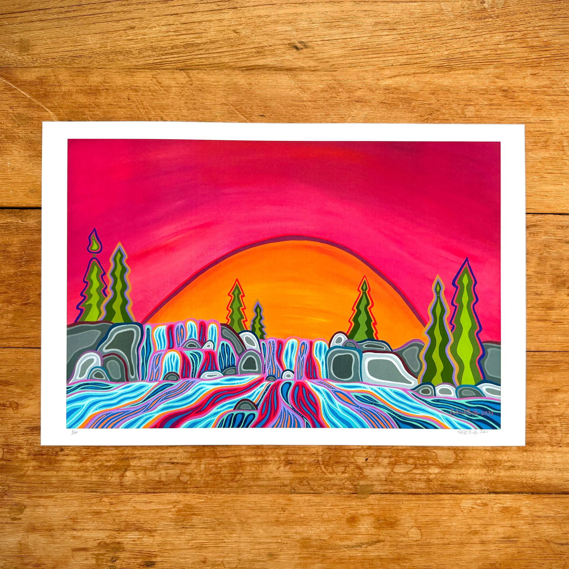 A print of Patrick Hunter's painting, Cascades, in Woodland Art style, against a wood grain backdrop. The print contains vibrant colours as cascading waterfalls run to the foreground, with trees, rocks surrounding. A large orange sun rests behind the cascades, against a bright sunset sky.