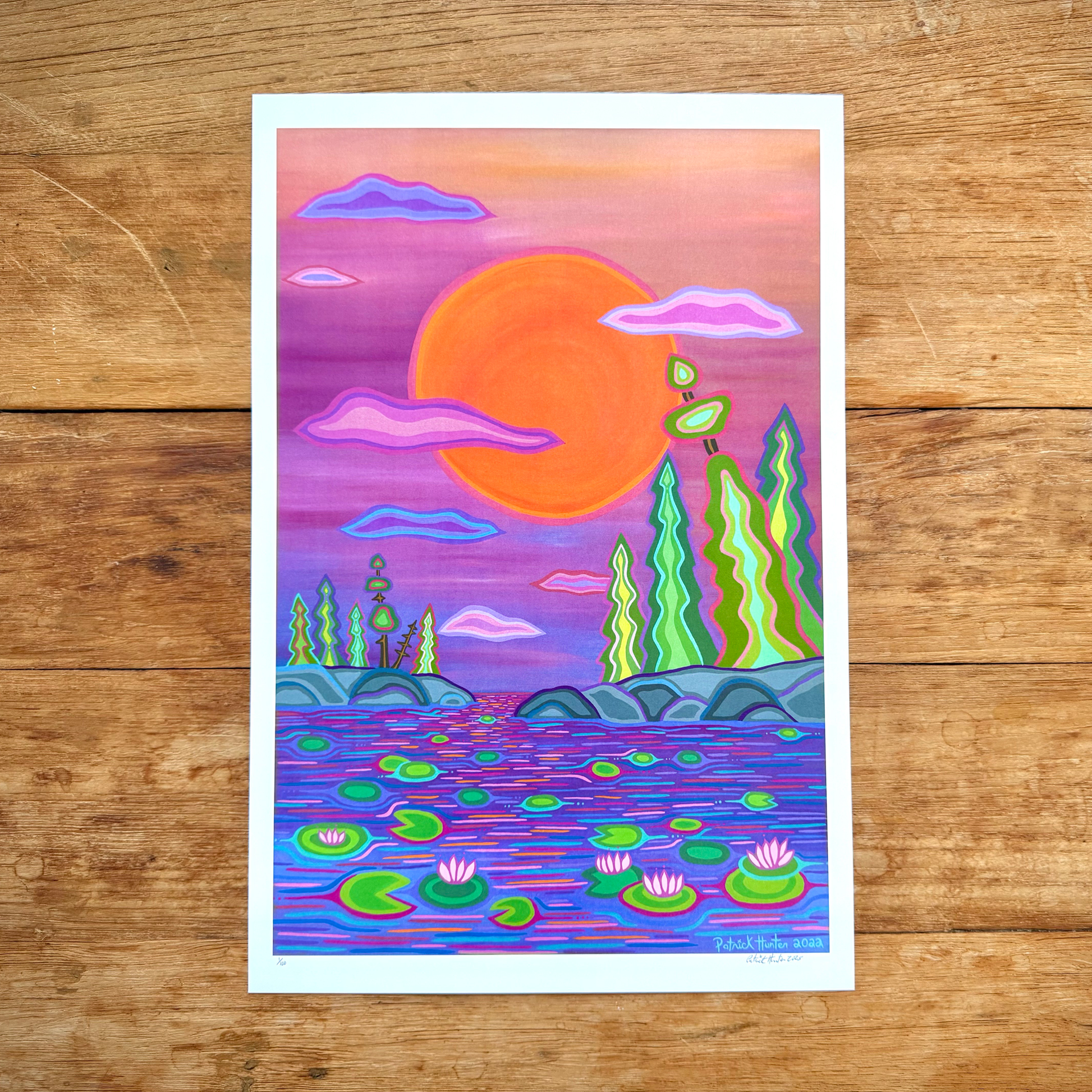 A print of Patrick Hunter's landscape painting, Sunset & Lillies, in woodland art style, of bright sun above water lily pond. The print rests on a wood grain background.