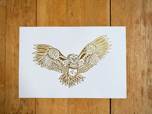 A print with gold leaf illustration of an owl with outstretched wings in Woodland art style on white archival paper against a wood grain background.