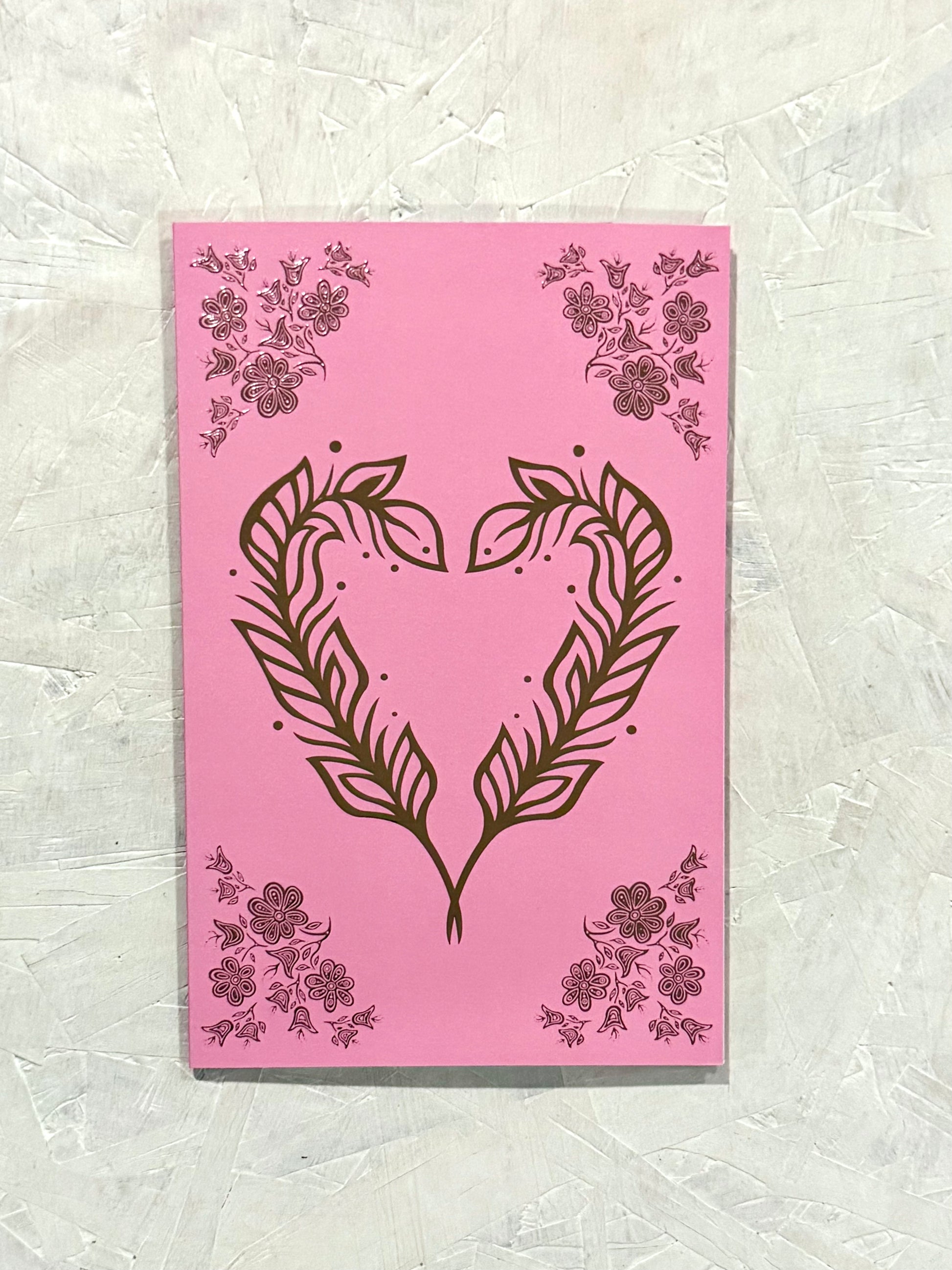Patrick's woodland art style design of two feathers in the shape of a heart with floral embellishments in the four corners of the card. The card contains raised rose gold foil elements, over a bubble gum pink soft touch laminate coating.