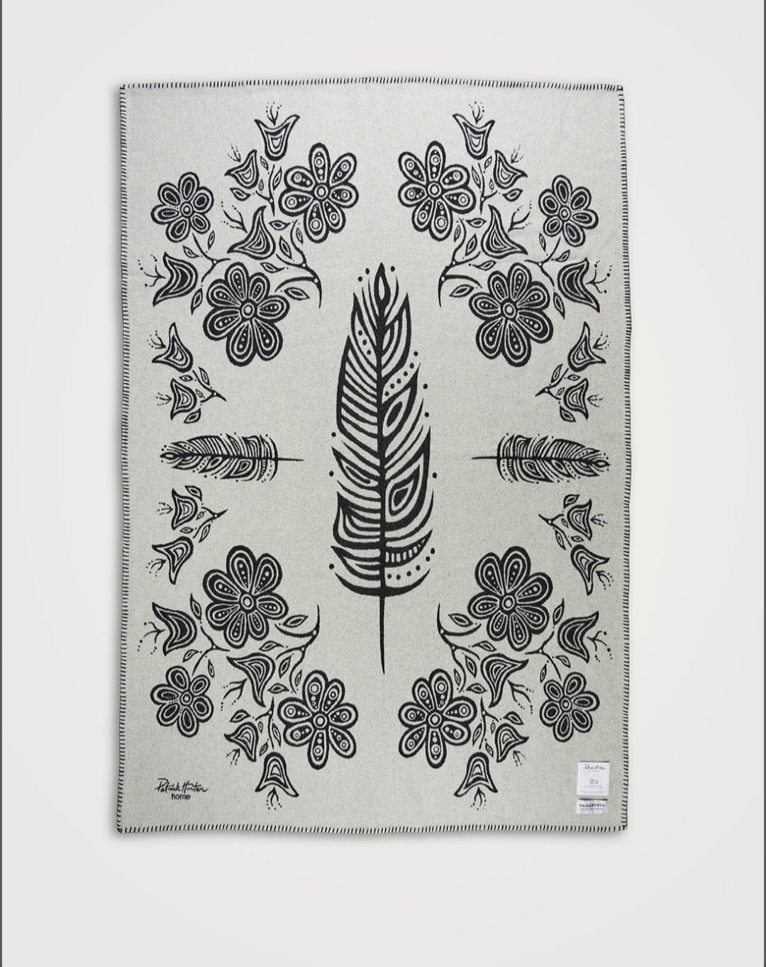 A white blanket with black woodland style line art of feathers and flowers, spread flat to show the full blanket design.
