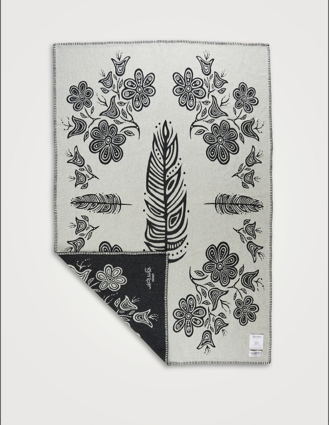 A white blanket with black woodland style line art of feathers and flowers, spread flat to show the full blanket design, with one corner lifted to show the reverse side is inverted — black with white art..