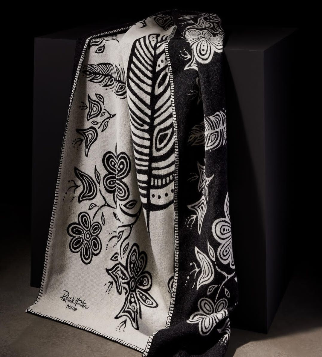 A black and white blanket with woodland style line art by Patrick Hunter is draped over a black surface.